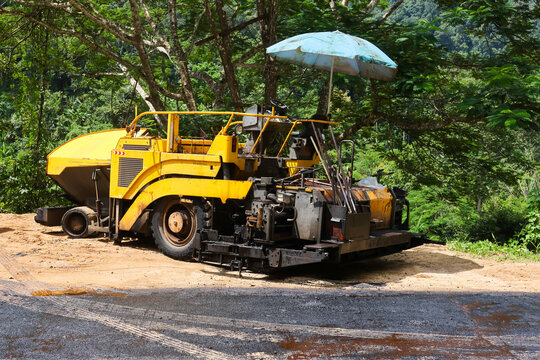 Yellow asphalt paving machine parked on forest road beneath sun umbrella, surrounded by tropical greenery and mountain terrain, with fresh gravel and construction work visible nearby