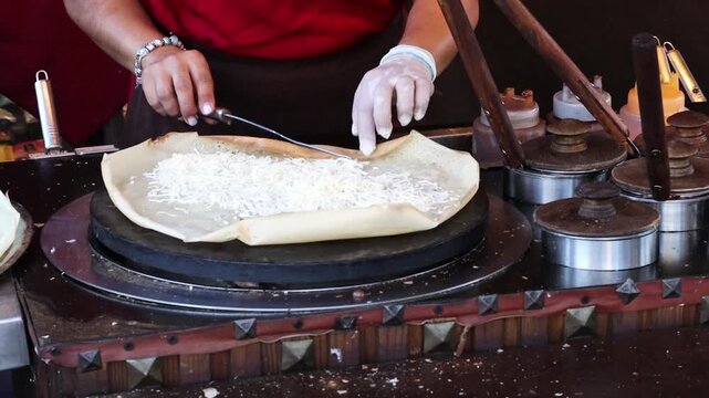 Large round flatbread cooking on a hot griddle at a busy outdoor food stall, with a vendor preparing traditional fair food using large pans and metal utensils during a festival.
