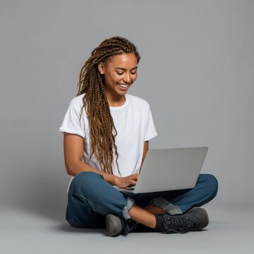 Happy young female student with braids typing on her computer while sitting on a grey background