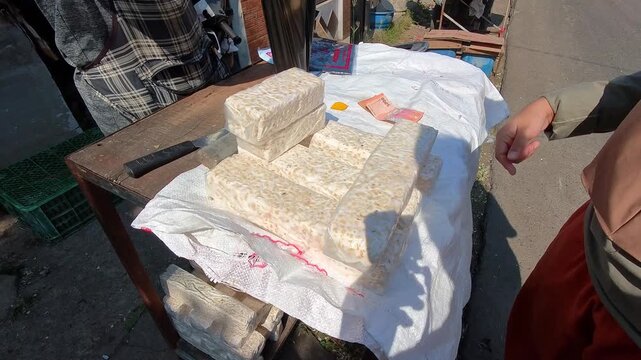 Video of Woman hands rest gently on white cloth selecting pile of tempeh. Stacked blocks sit on top of the fabric, neatly arranged. Person beside her adjusts a small blue item. Sunlight bathes the sce