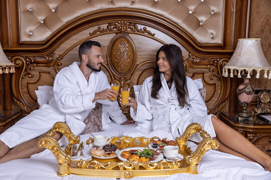 Couple in bathrobes toasting with orange juice over breakfast in a luxury hotel bed
