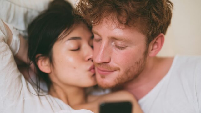 A wife of Asian decent is kissing her Caucasian ginger husband: men who are kissed by their partners are happier. Mixed-race couple is a bliss