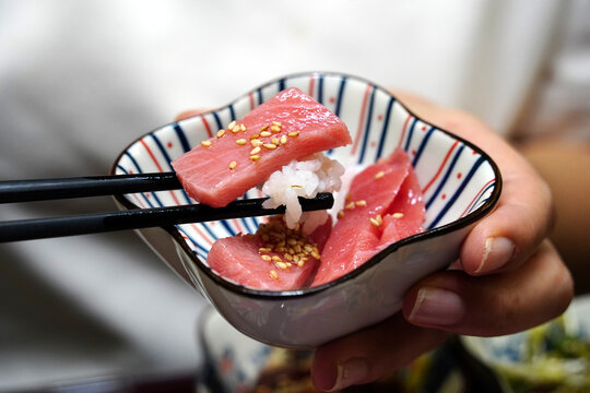 Chopsticks holding a fresh tuna sashimi slice with rice over a small bowl