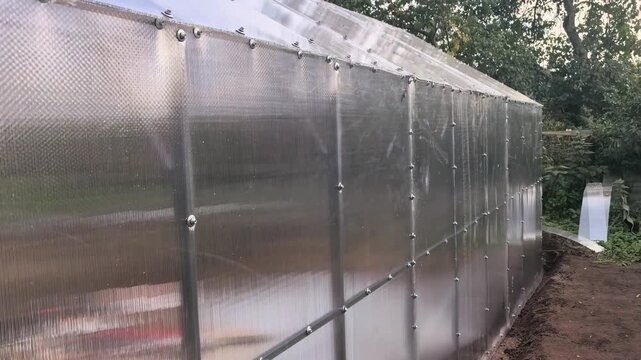 A greenhouse stands in a garden surrounded by trees and plants. The structure has clear panels and a metal frame. Sunlight shines on the scene.