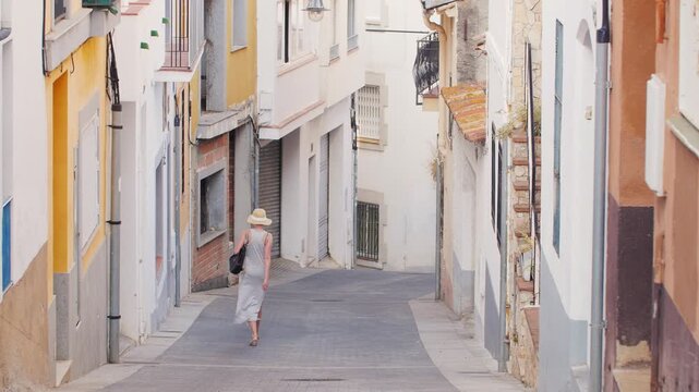 Woman in straw hat walking through narrow mediterranean street of old spanish town in summer.