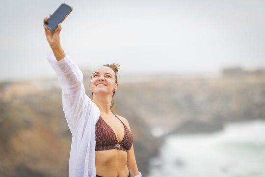 Woman takes selfie by the ocean during cloudy day near cliffs in coastal area