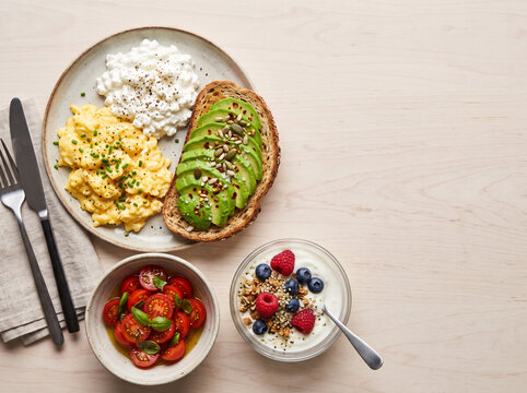 Breakfast spread with scrambled eggs, avocado toast, salad, and yogurt with fruit on a wooden table