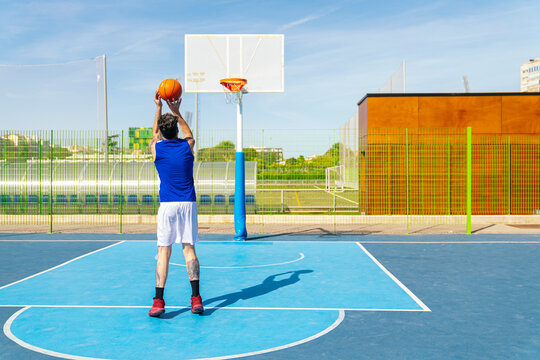 Male athlete aiming basketball for a shot on a vibrant blue outdoor court, practicing sports and enjoying leisure activity
