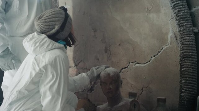 Scientist in protective suit inspects head closely, respirator and beanie visible, careful probe with gloved hand, mirror reflection shows cracked plaster, scattered vials and dust, tense forensic