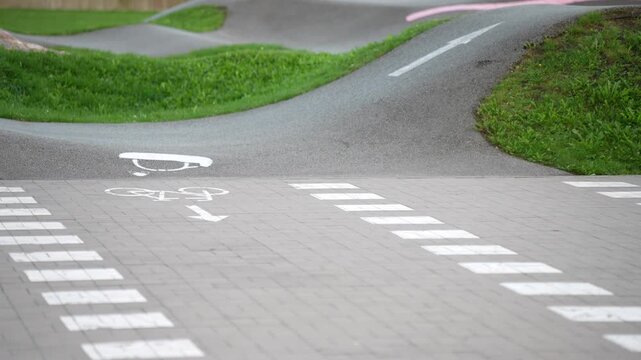 Scooter riders pass through a paved pump track with directional arrows and marked crossings, illustrating recreational riding routes and public active play infrastructure