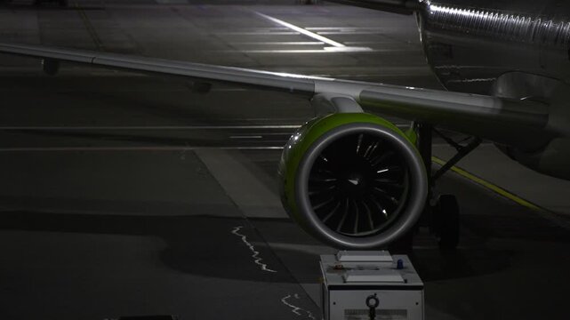 Fan blades turn inside an aircraft engine nacelle while the jet remains at its stand, showing turbofan components and adjacent ground support equipment on the apron