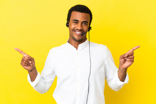 African American Telemarketer man working with a headset over isolated yellow background pointing finger to the laterals and happy