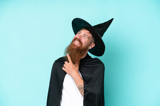 Young wizard in halloween isolated on blue background looking up while smiling