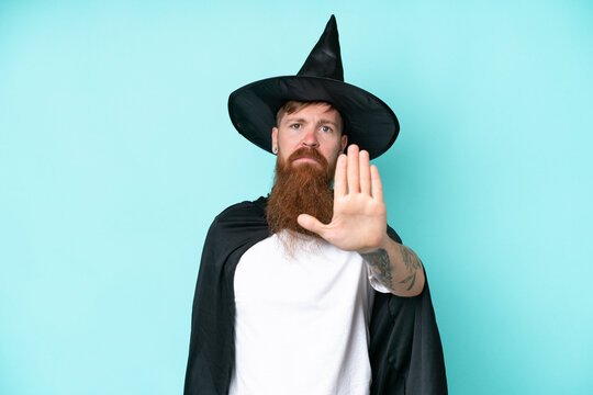 Young wizard in halloween isolated on blue background making stop gesture