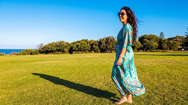 Woman in flowing turquoise kaftan walking on grass field by ocean, joyful summer portrait