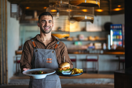 Friendly male waiter in apron serving delicious burger and fries to a guest in cafe