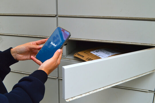Person holding smartphone displaying an online marketplace app while collecting a cardboard parcel from an automated self-service delivery locker