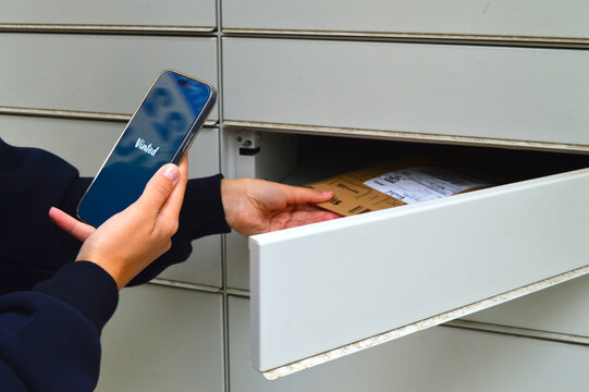 Person holding a smartphone displaying an online marketplace app while placing a cardboard parcel into an open automated postal locker outdoors