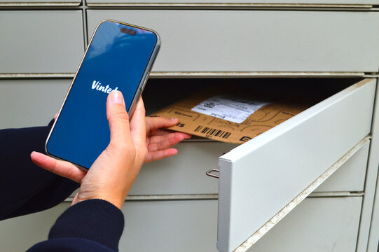 Woman using a smartphone with a secondhand shopping app open while inserting a cardboard delivery parcel into an outdoor parcel locker box