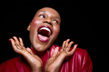 Obraz premium Joyful dark skinned African woman with bald head wearing red jacket laughs with open mouth and hands near face on black background. Concept for happiness, confidence, and beauty.