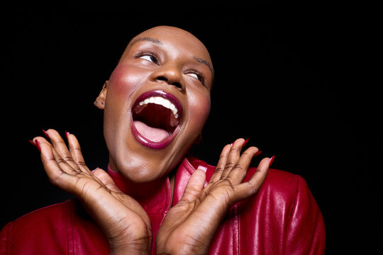 Joyful dark skinned African woman with bald head wearing red jacket laughs with open mouth and hands near face on black background. Concept for happiness, confidence, and beauty.