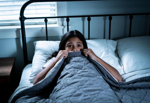 Scared young girl hiding under grey blanket in dark bedroom at night looking terrified with wide eyes blue moonlight