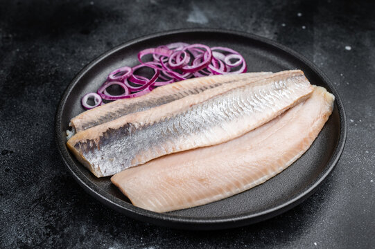 Pickled herring fillets cured in salt and brine served with onion and herbs. Traditional seafood on dark textured background.