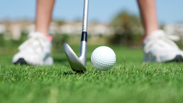 Close up of an iron club hitting a golf ball on a green grass course with a professional golfer's feet in the background, showcasing the perfect swing and impact during a sunny day