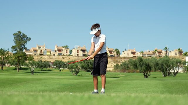 Young male golfer in a white visor executing a perfect swing with a golf club on a lush green course during a sunny day, with beautiful resort villas visible in the background