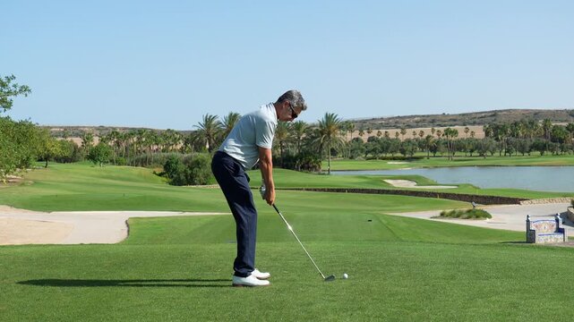 Professional golfer executing a powerful swing on a sunny day, driving the ball down the fairway of a luxury golf course with a lake and palm trees in the beautiful landscape