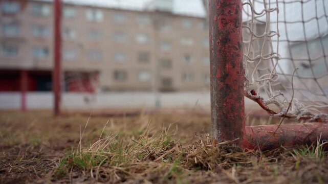 ground-level goalpost with grass foreground. low perspective shows rusted post, tangled net and muddy turf, stray ball rolling into frame, suburban apartment blocks in background, hopeful grassroots