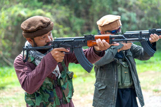 Two men in berets aiming AK-47 rifles in forested outdoor setting with focused expressions