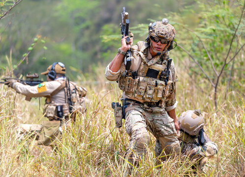 Soldier in tactical gear advancing through tall grass with rifle raised, forested background