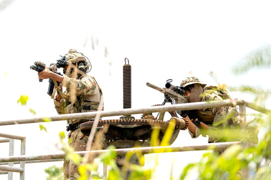 Two soldiers in camouflage gear aiming rifles from elevated metal platform amid bright foliage and overexposed sky