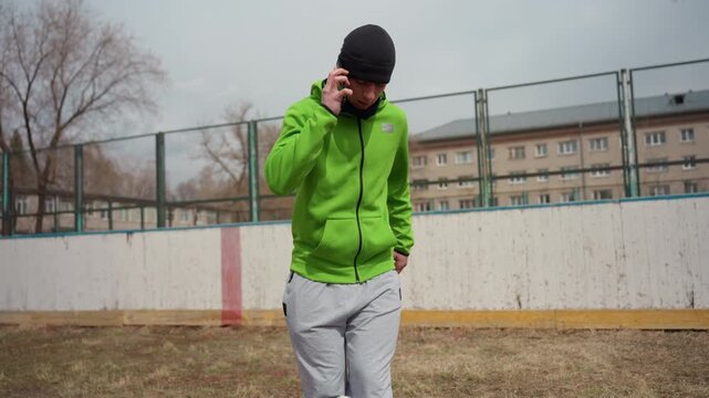 caucasian man walking on phone with soccer ball at feet, urban outdoor rink, green hoodie and grey pants, multitask moment blending conversation and casual juggling, relaxed training vibe