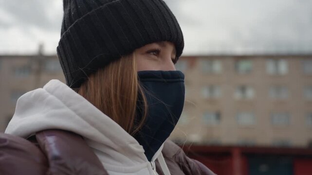 young woman wearing black beanie, standing outdoors in urban neighborhood, bundled in white hoodie and brown leather jacket, dark face mask covering lower face, neutral expression watching street,