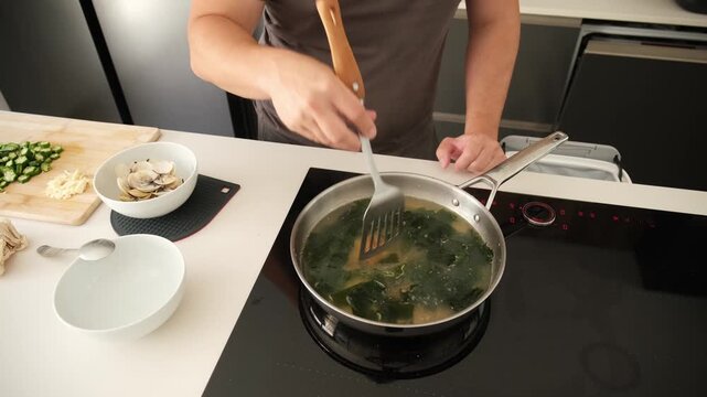 Man stirring a hot pot of miso soup with seaweed and clams on an induction stove