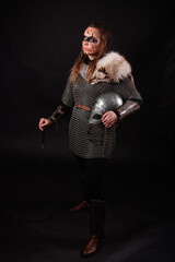 Obraz premium A young Viking woman in fur-trimmed chainmail and war paint, holding a sword, poses against a dark studio background.