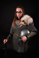 Obraz premium A young Viking woman in fur-trimmed chainmail and war paint, holding a sword, poses against a dark studio background.