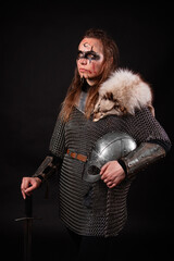 Obraz premium A young Viking woman in fur-trimmed chainmail and war paint, holding a sword, poses against a dark studio background.