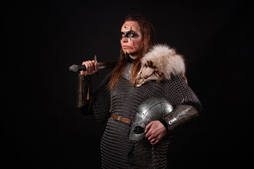 Obraz premium A young Viking woman in fur-trimmed chainmail and war paint, holding a sword, poses against a dark studio background.