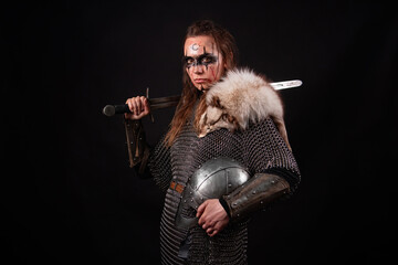 Obraz premium A young Viking woman in fur-trimmed chainmail and war paint, holding a sword, poses against a dark studio background.