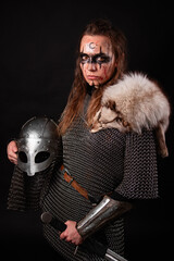 Obraz premium A young Viking woman in fur-trimmed chainmail and war paint, holding a sword, poses against a dark studio background.