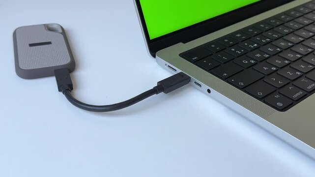 Male hand plugging external SSD into laptop USB-C port with green screen display, white table close up