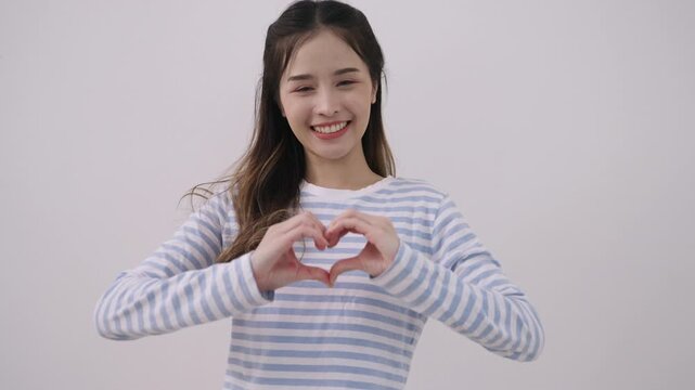 Happy, beautiful young Asian woman standing, smiling, with arms crossed and posing with a heart shape with her hands while looking at the camera with a fun and cheerful white studio background.
