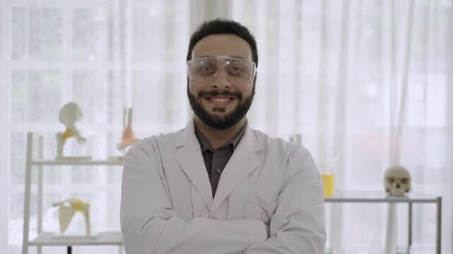 Portrait of happy handsome Middle Eastern man scientist wearing white lab coat and safety goggles, smiling and standing with arms crossed, thumb up, while looking at the camera in science laboratory. 