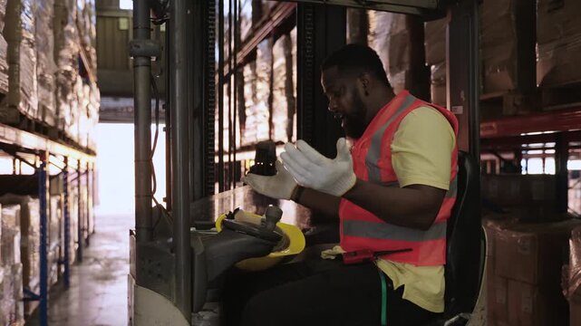 Unhappy African American male forklift driver sits in a vehicle, removes his yellow helmet, and talks and grumbles to himself, expressing his frustration with working at distribution warehouse center.