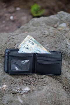 An open black leather wallet filled with Indonesian Rupiah banknotes lying on a rocky ground texture, illustrating concepts of lost wallet, personal finance, and limited