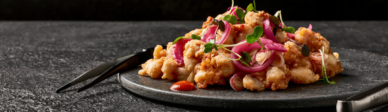 Gourmet fried chitterlings plated with pickled red onions and microgreens on a dark slate plate, served with chili sauce and chopsticks, restaurant food presentation, appetizer or main course