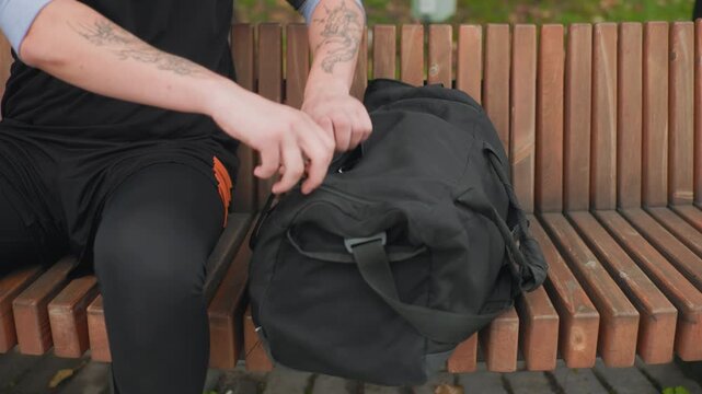 white male unpacking black duffel bag on wooden park bench pulling out blue hand wraps and gloves tattoos visible on forearms preparing for boxing workout boxer checks wraps runner grabs water trainer